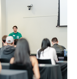 A Heterodox Research Society member in a green society shirt addressing a seated audience in a lecture room