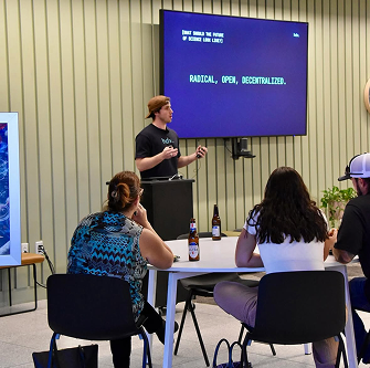 A speaker presenting to attendees at tables in front of a screen reading Radical, Open, Decentralized